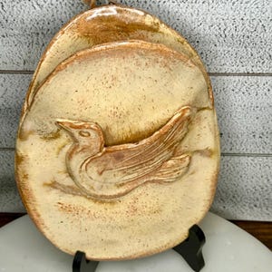 May include: A ceramic wall hanging with a bird design. The oval-shaped piece features a raised relief of a bird in shades of tan and brown. The background is a lighter tan color with darker speckles. The piece is mounted on a black stand.