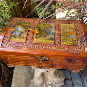 May include: A rectangular wooden box with a polished finish, featuring three inset panels on the lid that display colorful landscape illustrations. The box is decorated with intricate carved scrollwork borders and includes a central metal latch.