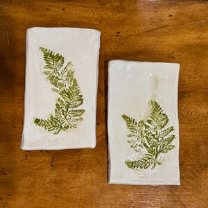 May include: Two rectangular ceramic plates with a glossy off-white finish. Each plate features a single green fern leaf imprint centered on the surface. The edges of the plates are slightly irregular, and a small maker's mark is visible in the corner.