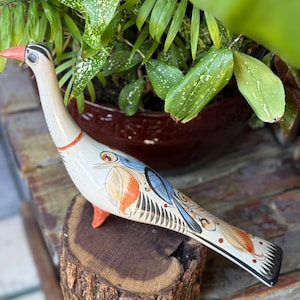 May include: A hand-painted, decorative bird figurine. The bird has a cream-colored body with orange, blue, and black accents. It has a long neck, a red beak, and is perched on a wooden base. The bird is a unique piece of home decor.