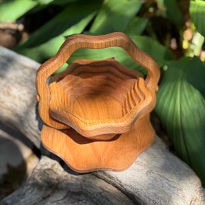 May include: A wooden basket with a handle, featuring a layered design. The basket is made of light brown wood and has a scalloped edge. The handle is curved, and the basket sits on a matching base. The background includes green foliage.