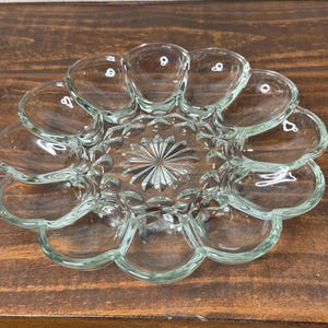 May include: Clear glass egg serving dish with a scalloped edge and a central starburst design. The dish has 12 individual egg-shaped compartments. The dish is made of clear glass and sits on a dark wooden surface.