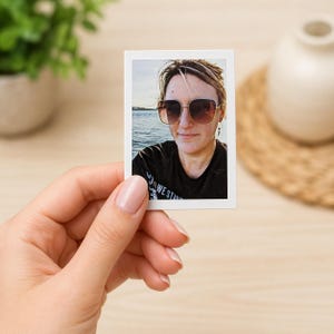 May include: A small, rectangular photograph held by a hand. The photo features a person wearing sunglasses and a black shirt with the text "WE STAND". The background shows water and a bright sky.