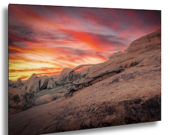 May include: A landscape photograph of a desert scene featuring a natural rock arch under a vibrant sunset sky. The sky is filled with streaks of red, orange, and yellow. The rock formations are a warm, reddish-brown color.
