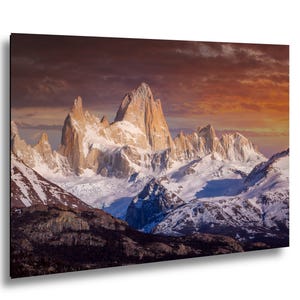 May include: A landscape photograph of a mountain range with snow-covered peaks under a dramatic sunset sky. The mountains have a rugged, textured appearance, with snow and ice clinging to their slopes. The sky is a mix of orange, red, and purple hues.
