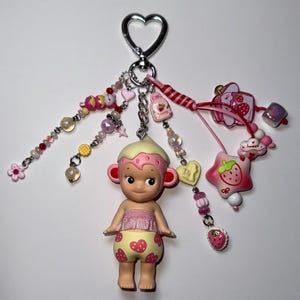 Puede incluir: Un llavero caprichoso con una peque&ntilde;a mu&ntilde;eca con un atuendo con tem&aacute;tica de fresas y un tocado color crema. El llavero incluye varios dijes, como cuentas, corazones y una estrella con forma de fresa, en tonos de rosa, rojo y amarillo.