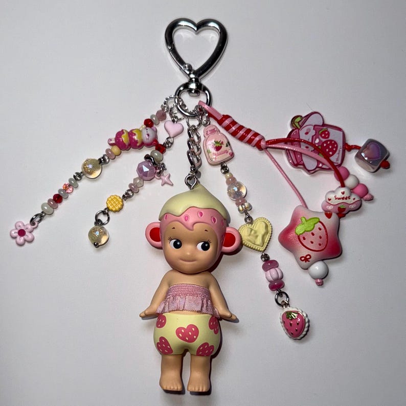 Puede incluir: Un llavero caprichoso con una peque&ntilde;a mu&ntilde;eca con un atuendo con tem&aacute;tica de fresas y un tocado color crema. El llavero incluye varios dijes, como cuentas, corazones y una estrella con forma de fresa, en tonos de rosa, rojo y amarillo.