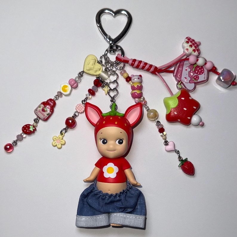 Puede incluir: Un llavero caprichoso con una mu&ntilde;eca vestida de fresa, con una camiseta roja con una flor blanca y pantalones cortos de mezclilla. El llavero incluye varios amuletos como una estrella, corazones y cuentas, todo con tem&aacute;tica de fresa.