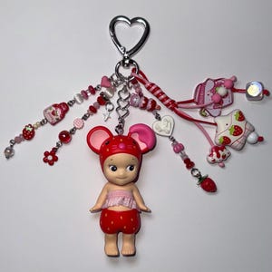 Puede incluir: Un llavero caprichoso con una mu&ntilde;eca de rat&oacute;n fresa. El llavero incluye un cierre plateado en forma de coraz&oacute;n y varios dijes rosas y rojos, incluyendo cuentas, corazones y decoraciones con tem&aacute;tica de fresas. La mu&ntilde;eca lleva un atuendo rojo con un estampado de fresas.