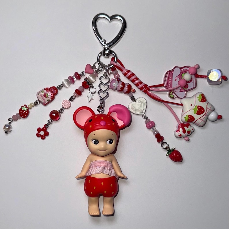 Puede incluir: Un llavero caprichoso con una mu&ntilde;eca de rat&oacute;n fresa. El llavero incluye un cierre plateado en forma de coraz&oacute;n y varios dijes rosas y rojos, incluyendo cuentas, corazones y decoraciones con tem&aacute;tica de fresas. La mu&ntilde;eca lleva un atuendo rojo con un estampado de fresas.