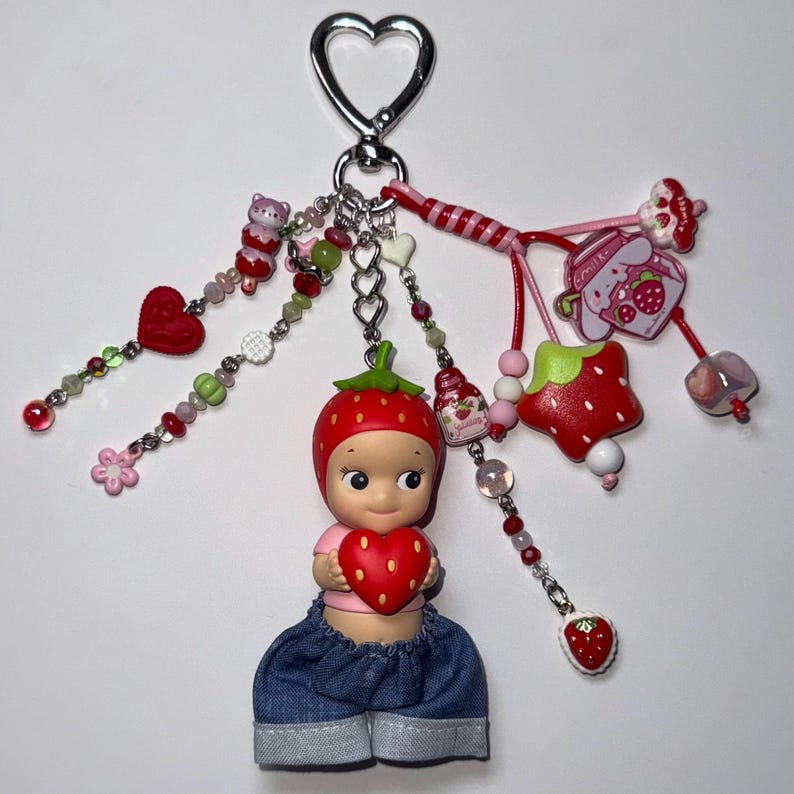 Puede incluir: Un llavero caprichoso con una mu&ntilde;eca que lleva un sombrero de fresa y pantalones cortos de mezclilla, sosteniendo un coraz&oacute;n rojo. El llavero incluye varios dijes como una fresa, un oso y cuentas, conectados a un cierre plateado en forma de coraz&oacute;n.