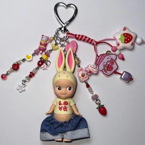 Puede incluir: Un llavero caprichoso con una mu&ntilde;eca con orejas de conejo, que lleva una camiseta amarilla con "LOVE" y una fresa, y pantalones cortos de mezclilla. El llavero incluye varios dijes: fresas, estrellas, corazones y cuentas, todos unidos a un cierre plateado en forma de coraz&oacute;n.