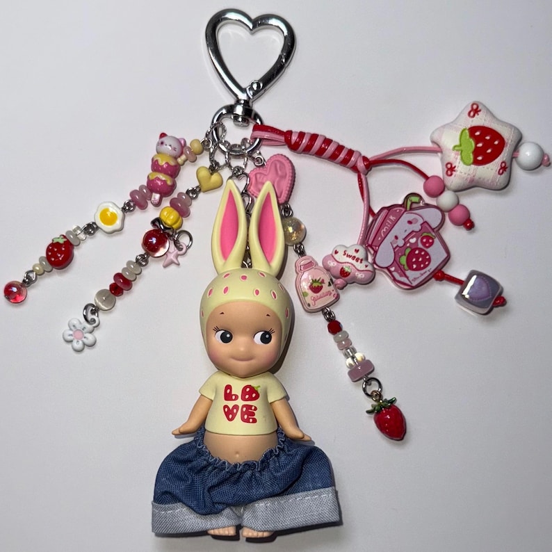 Puede incluir: Un llavero caprichoso con una mu&ntilde;eca con orejas de conejo, que lleva una camiseta amarilla con "LOVE" y una fresa, y pantalones cortos de mezclilla. El llavero incluye varios dijes: fresas, estrellas, corazones y cuentas, todos unidos a un cierre plateado en forma de coraz&oacute;n.
