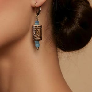 May include: A drop earring featuring a cylindrical bronze-toned metal bead with an intricate embossed pattern, flanked by two small, round, light blue translucent beads. The earring is suspended from a simple bronze-toned metal hook wire.