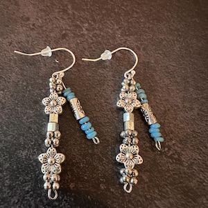 May include: A pair of dangle earrings featuring silver-toned metal flower charms, small round beads, and cylindrical textured spacers. Each earring includes a secondary strand accented with light blue seed beads and a silver-toned metal tube bead.