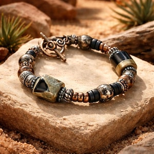 May include: A handcrafted bracelet featuring a mix of metallic beads in silver, bronze, and black. The bracelet includes a large, square-shaped bead with a dark stone inlay and a toggle clasp. The design has an earthy, bohemian aesthetic.