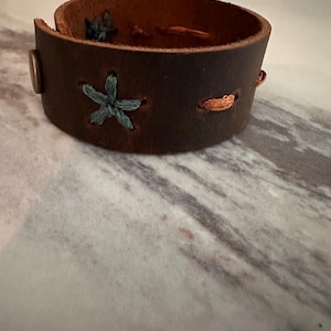 May include: A brown leather cuff bracelet with a stitched star design in teal thread. The bracelet has a metal button closure and orange stitching details. The interior of the bracelet is a lighter brown color.