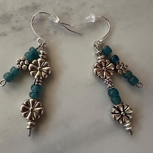 May include: A pair of dangle earrings with silver-tone metal and teal beads. Each earring features a flower-shaped charm, small beaded clusters, and a series of teal beads. The earrings have silver-tone ear wires.