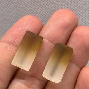 May include: Two rectangular, translucent glass pieces with a gradient effect, transitioning from a light brown at the top to a clear, frosted appearance at the bottom. The pieces are held in a person's hand, showcasing their shape and colour.