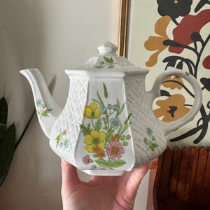 May include: White ceramic teapot with a floral design. The teapot features yellow, pink, and blue flowers, a lattice pattern, and a lid. A classic piece for serving tea, perfect for any kitchen.