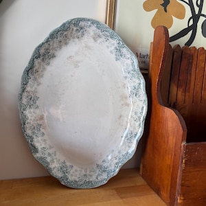 May include: An oval, antique serving platter with a scalloped edge. The rim features a floral pattern in shades of blue and white. The centre of the platter is white with subtle floral accents. The platter is displayed on a wooden surface.