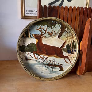 May include: A vintage round metal tray with a winter scene depicting a running deer, trees, and snow. The tray has a gold-coloured rim and a painted illustration. The artist's name, James L. Artis, is visible on the tray.