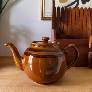 May include: A glazed ceramic teapot featuring a rounded body with a glossy finish in shades of warm amber and dark brown. The teapot includes a matching lid with a small knob, a curved spout, and a sturdy handle, showcasing horizontal bands.