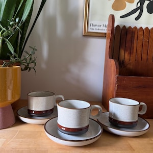 May include: Three ceramic coffee cups and saucers. The cups are speckled beige with a brown band and rim. The saucers have a matching brown rim. The set is displayed on a wooden surface, with plants and a framed print in the background.
