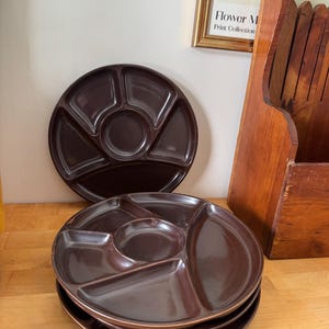May include: Four dark brown divided serving platters, each with a central circular section and four surrounding compartments. One platter is placed above the stack. The platters are on a wooden surface.