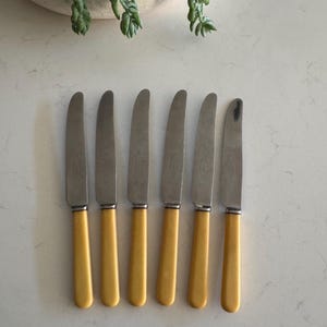 May include: A collection of seven vintage knives, each with a stainless steel blade and a yellow handle. The knives are laid out in a row. The handles are a pale yellow, and the blades are silver. The knives are on a white surface.