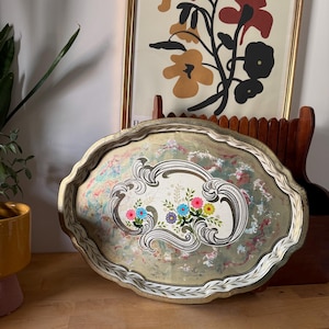 May include: An oval metal serving tray with a scalloped edge, featuring a central white panel decorated with a colourful floral bouquet of blue, pink, and yellow flowers, surrounded by an ornate grey scrollwork border and a distressed green finish.