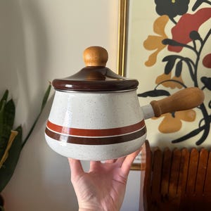 May include: A vintage white speckled enamel saucepan with a brown lid and handle. The pot has two brown and red stripes. The lid has a wooden knob. A classic kitchenware item, perfect for cooking.