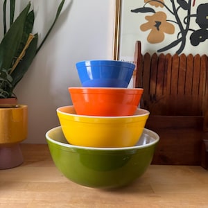 May include: Four brightly coloured mixing bowls stacked on top of each other. The bowls are in shades of green, yellow, orange, and blue. The largest bowl is green, and the smallest is blue. They are on a wooden surface.
