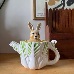 Peut inclure: Une théière en céramique blanche avec un motif de feuilles vertes et une figurine de lapin qui regarde. La théière a une poignée et un bec courbés. Le lapin est beige et a de longues oreilles. La théière est sur une surface en bois.