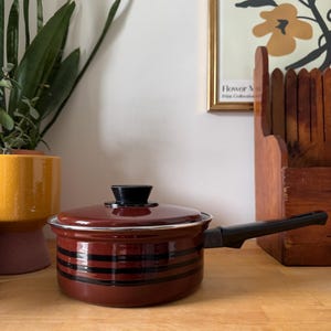 Peut inclure: Une casserole marron avec des rayures horizontales noires et une poignée noire. Le pot a un couvercle assorti avec un bouton noir. Le pot est posé sur une surface en bois.