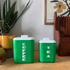 May include: Two green and white kitchen canisters. The larger canister has the word "COFFEE" in white vertical text. The smaller canister has the word "TEA" in white vertical text. Both canisters have white lids.