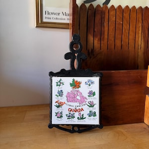 Peut inclure: Un dessous de plat en métal noir avec un carreau blanc central orné des mots "Floral Emblems Canada" et d'illustrations de diverses fleurs. Le dessous de plat a une poignée décorative et une bordure détaillée.
