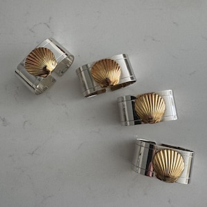 May include: Four silver napkin rings, each featuring a gold seashell design. The rings are rectangular, with a gold seashell detail in the centre. They are set against a light grey background.