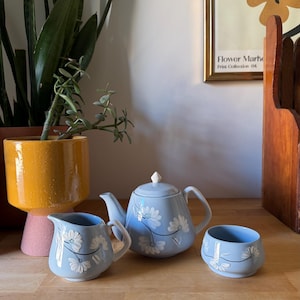 May include: A pale blue ceramic tea set decorated with white floral patterns. The set comprises a teapot, a milk jug, and a sugar bowl. The teapot has a lid with a white knob. The floral design features white flowers with black stems. The set is on a wooden surface.