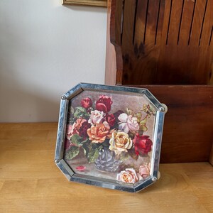 Peut inclure: Un cadre octogonal argenté contenant une image d'une nature morte florale. L'œuvre représente un vase débordant de roses rouges, roses et oranges. Le cadre est en métal argenté et a une esthétique vintage.