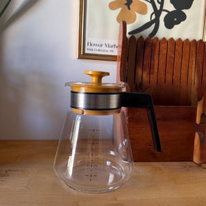 Peut inclure: Une carafe à café en verre transparent dotée d'un couvercle jaune moutarde avec un bouton central, d'une bande en acier inoxydable autour du col et d'une poignée en plastique noir. Le corps en verre présente des graduations noires de 4 à 12.