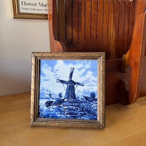 Peut inclure: Un carreau encadré représentant un paysage de moulins à vent bleu et blanc. L'œuvre d'art représente un paysage néerlandais avec des moulins à vent, de l'eau et un ciel nuageux. Le cadre est doré avec un motif décoratif.