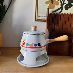 Peut inclure: Pot à fondue blanc avec une poignée et un couvercle en bois. Le pot porte un motif rouge, orange et bleu avec le mot "fondue" sur le côté. Il repose sur un support blanc avec une base circulaire. Un article de cuisine vintage.