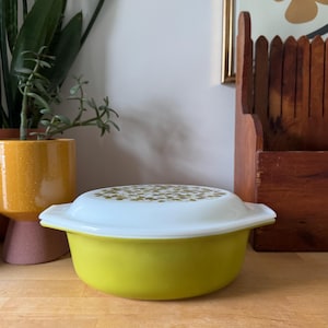 May include: A vintage yellow and white casserole dish with a floral pattern on the lid. The dish has a rounded shape and a white lid with a gold floral design. The dish is on a wooden surface.