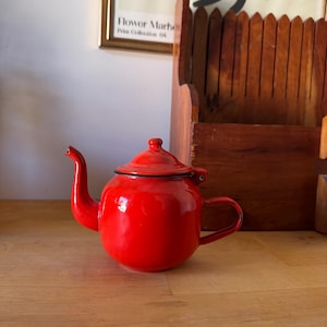 May include: A bright red enamel teapot with a matching lid, a curved spout, and a side handle. The teapot features a smooth, glossy finish and a black rim around the top opening where the lid rests. The design is simple, rounded, and functional.