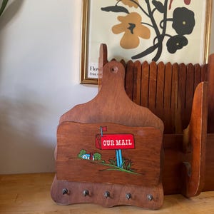 May include: A wooden wall-mounted mail organiser featuring a cutout shape with a central pocket. The front of the pocket displays a painted graphic of a red mailbox with the text OUR MAIL, a small house, and green grass. Five metal hooks are attached.