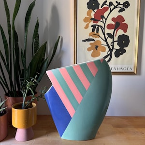 May include: A large, colourful ceramic vase with a geometric design. The vase features sections of blue, pink, and green. A framed floral print hangs in the background, and potted plants are on the left.