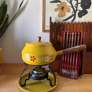 May include: A yellow enamel fondue pot with a floral pattern, a wooden handle, and a matching lid. It sits on a black metal stand with a burner and a yellow base plate. Beside it is a red plastic case containing six metal fondue forks with wooden handles.