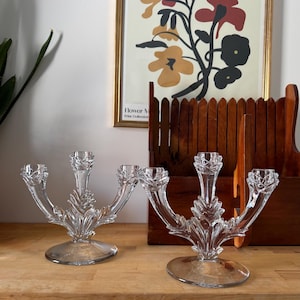 Peut inclure: Deux candélabres en verre transparent, chacun avec trois bras. Les candélabres présentent un motif de feuilles sur les tiges et une base ronde. Ils sont posés sur une surface en bois.