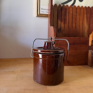 May include: A small, brown ceramic jar with a lid and a metal handle. The jar is cylindrical and sits on a wooden surface. The background includes a wooden rack and a framed print.